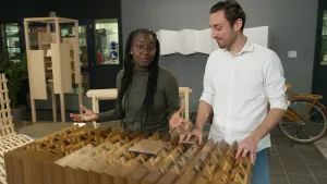 Een foto van presentator Tirsa die in een school staat. Ze staat achter iets van hout. Het houten ding heeft een ingewikkelde vorm. Naast haar staat een man. Tirsa is iets aan het vertellen. 