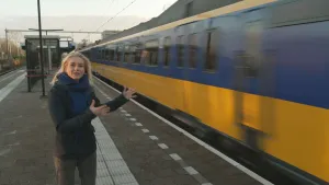 Een foto van presentator Janouk. Ze staat op een treinperron en vertelt iets. Naast haar raast een trein voorbij. 