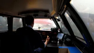 Een foto van een treinmachinist in de bestuurdersruimte van de trein. De machinist kijkt voor zich door de ruit en links en rechts in de ruimte zijn ook ruiten te zien. Hij zit voor knopjes, metertjes, een lichtje, een hendel, een tablet en een camera.