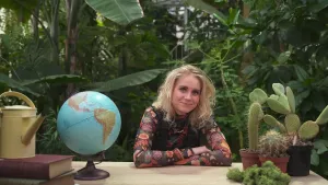 Een foto van Janouk achter een tafel met haar armen over elkaar. Rechts op tafel ligt een pluk groen mos voor drie cactussen met scherpe naalden en een lange of bolle vorm. Links staat een wereldbol op tafel en achter Janouk staan groene planten.