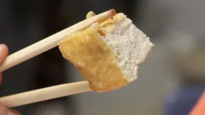 Een foto van een blokje tofu dat met twee houten eetstokjes voor de camera gehouden wordt. Het blokje heeft een goudgele korst en een witte binnenkant. Tofu wordt gemaakt van blokjes soja en kan daarom gegeten worden bij een vegetarische maaltijd. 