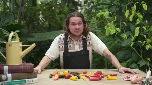Een foto van Pascal met achter zich groene planten en voor zich op tafel pepers die rood, geel of groen van kleur zijn. Links op de tafel staat een gele gieter op een stapel van drie boeken en rechts op tafel liggen een klein tuinharkje en tuinschepje. 
