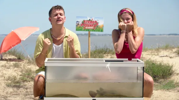 Een foto van Emma en Jurre die op een strand zitten. Voor hen staat een glazen bak met daarin een Gemarmerde Sidderrog. Ze kijken allebei heel bang.