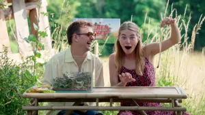 Een foto van Jurre en Emma die buiten aan een tafel zitten. Op de tafel staat een glazen bak met planten erin. Emma houdt een slangenhuid in de lucht en kijkt verbaasd. Jurre lacht naar haar.