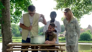 Een foto van Jurre en Emma en twee andere mensen. Ze staan buiten, bij een tafel. Op de tafel staat een glazen bak met water en een Dwergmeerval erin. Jurre twijfelt of hij zijn hand in de bak gaat steken.