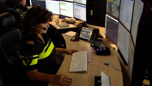 Een foto van een politieagent achter een bureau die zes beelschermen voor zich heeft staan in rijen van drie. De politieagent heeft een hoofdtelefoon met microfoon op om de mensen die het noodnummer 1 1 2 bellen op de grote telefoon ernaast te spreken.