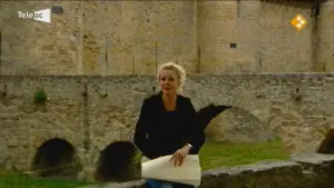 Een foto van een vrouw die op een stenen muurtje zit. Ze heeft een papier in haar handen. Achter haar zie je een oude stenen brug met daarachter een kasteelmuur.