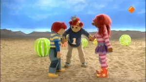 Een foto van Koning Koos, Sassa en Toto. Ze staan op een strand. Koning Koos houdt de handen vast van Sassa en Toto. Op de achtergrond zie je enkele strandballen in het zand.