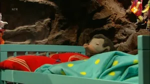 Een foto van Moffel bij zijn bed. Het bed heeft rode kussens en een groene deken met gele stippen erop.