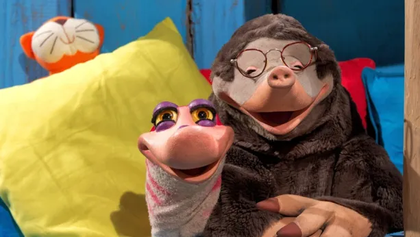 Een foto van Moffel en Piertje. Ze kijken je lachend aan. Op de achtergrond zie je enkele felgekleurde kussens. 