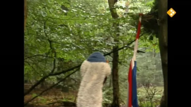 Een foto van Arie in het bos. Op de achtergrond zie je de Nederlandse vlag op een stok in de grond staan. 