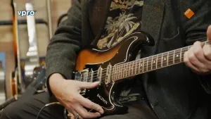Een foto van een man met een elektrische gitaar op schoot. Hij speelt op die gitaar.  Zijn gezicht is buiten beeld.