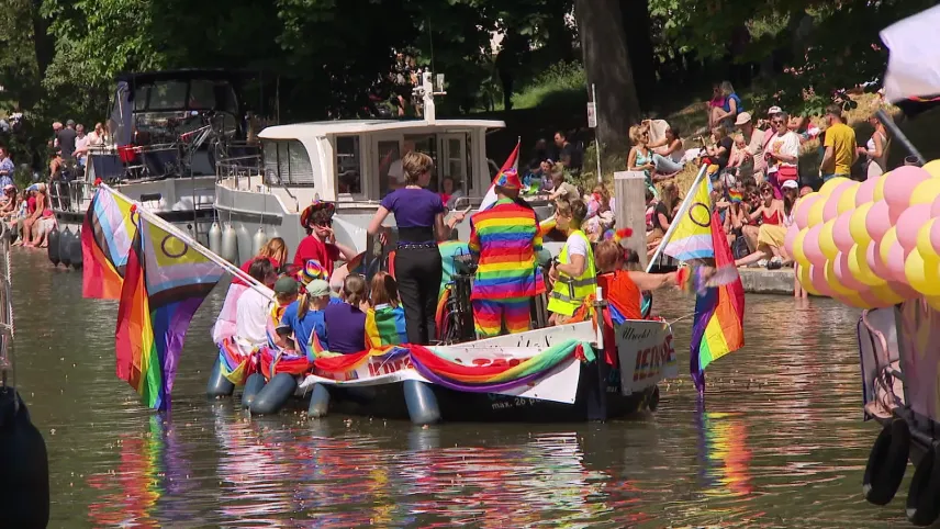 Een foto van de viering van Pride-maand voor LHBTQA-plussers. Er is een boot te zien met allemaal mensen erin die vlaggen met regenboogkleuren vasthouden. Aan de kant in het gras zitten mensen naar de optocht, waar de boot een onderdeel van is, te kijken.