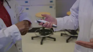 Een foto van een klein apparaat met een rood knopje, waarmee een prik zonder naald gezet kan worden. De medewerker van de Universiteit Twente die het apparaat vastheeft, heeft een prik gezet op de hand van de presentator, die over haar eigen hand wrijft.