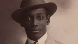 Een oude zwart-wit foto van het gezicht van de Surinaamse held Anton de Kom. Hij draagt een donkere hoed. Op de foto is ook de bovenkant van zijn kleding nog te zien. Hij draagt een lichtgekleurd pak met een witte blouse eronder en heeft een stropdas om. 