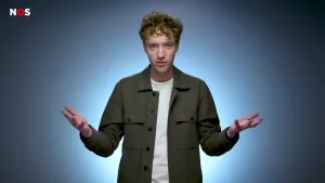 Een foto van een presentator die zijn handen met de handpalmen omhoog naast zich in de lucht houdt. Hij heeft een donkergrijze blouse met een wit t-shirt eronder aan. De presentator staat voor een blauwgrijze achtergrond met wit licht om hem heen. 