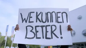 Een foto van de handen van een persoon die een wit protestbord vasthebben. Op het protestbord is met zwarte letters de tekst "We Kunnen Beter!" geschilderd. De persoon staat buiten, want er zijn bomen, een gebouw en een blauw met witte lucht te zien.