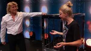Een foto van de bekende illusionist Hans Klok die zijn arm op een grote, zwarte kooi heeft liggen. Een illusionist goochelt met grotere voorwerpen en heeft assistenten in zijn spannende show. Rechts staat presentator Janouk, die met Hans praat..