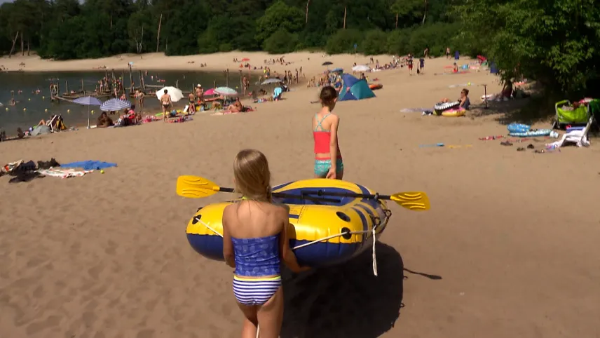 Een foto van twee meisjes die op het strand een blauw met geel gekleurde rubberboot dragen, waar de peddels van de boot opliggen. De meisjes lopen van de camera weg en hun rug is te zien op de foto. Op het strand liggen verderop mensen in de zon.