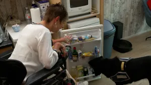 Een foto van een vrouw die in haar rolstoel in de keuken zit. Ze heeft de koelkast openstaan. Naast de koelkast staat haar zwarte hulphond, een hond die speciaal getraind is om haar te helpen. De hond, die Noemie heet, pakt een flesje uit de koelkast.
