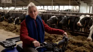 Een foto van een koeienstal waar koeien hooi aan het eten zijn op de achtergrond. Het zijn allemaal wit met zwart gekleurde koeien. Op de voorgrond zit presentator Janouk op een quad, dat is een vierwielige brommer. Janouk kijkt naar de camera.