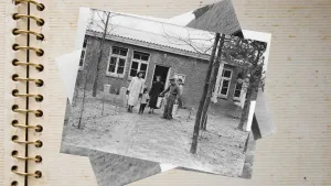 Een oude zwart-wit foto van een pand van woonoord Lunetten in Vught. Op het pad voor het pand staan vijf Molukse mensen. Twee vrouwen, een kind, een medewerker met een pet op en in zijn handen twee langwerpige emmers en erachter een man met een tuinhark.