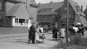 Een zwart-witte foto van mensen die over een straat lopen. Een stel loopt met een kinderwagen met een klein kind erin. Andere mensen lopen met grotere karren waarin ze oudere mensen en spullen vervoeren. De straat loopt door een wijk met grote huizen.