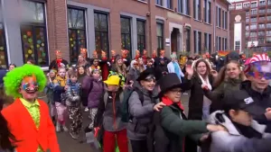 Een foto met feestvierende mensen die carnaval vieren. Vooraan lopen kinderen een polonaise. De meeste kinderen hebben alleen een carnavalshoed op. Links van de polonaise staat een kind met een groene pruik, feestneus, rood jasje en gele blouse. 