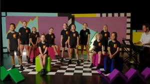 Een foto van elf kinderen in een zwart t-shirt met een rood-geel Kinderen voor Kinderen logo erop. Op de zwart-wit geblokte vloer staan zes kinderen en vijf kinderen zitten op een gekleurde kubus.  Zangcoach Michiel staat achter een elektrische piano.