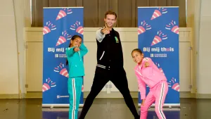 Een foto van dansleraar Olivier en de kinderen Gaby en Nova die naar de camera wijzen. Daarachter staan twee blauwe doeken op een standaard. Op de doeken staan de teksten "Bij mij thuis" en "Kinderen voor Kinderen" tussen roze met witte confetti kanonnen.