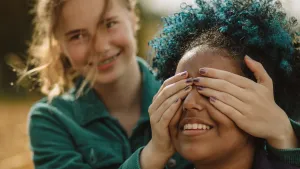 Foto van een meisje die de handen voor de ogen van een ander meisje houdt en beiden lachen ze.