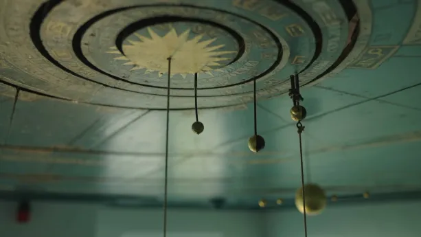 Een foto van een planetarium op een plafond. Een planetarium is een apparaat waarmee de sterrenhemel wordt getoond. Op het plafond zijn cirkels met een zon in het midden. Daar hangen balletjes vanaf die de zon en planeten zijn.