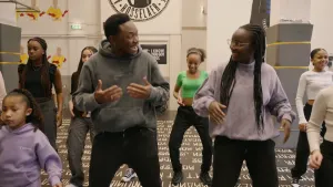 Een foto van een groep mensen die in twee rijen aan het dansen zijn. In het midden staan presentator Tirsa en een man die iets aan haar uitlegt. Er staan teksten op de vloer van de kamer. Twee pilaren in de ruimte hebben een dun stootkussen om zich heen.