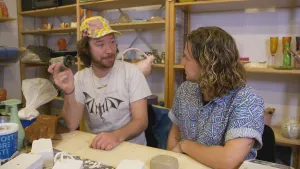 Een foto van presentator Pascal en een andere man. Zij zitten achter een tafel en praten met elkaar. De man heeft een voorwerp in zijn hand, een kleien versie van een camera. Achter hen staat een kast met kleien beeldjes.