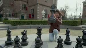 Een foto van presentator Serah die op de grond geknield zit bij een groot schaakbord. Voor haar staan de zwarte schaakstukken. Ze houdt een schaakstuk in haar handen omhoog. Achter haar zijn strak geknipte struiken te zien en een kasteel. 