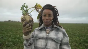 Een foto van presentator Eva, die buiten op een veld met planten staat. In haar hand heeft ze een suikerbiet. Dit is een grote knol met wat stengels met blaadjes eraan. 