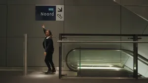 Een foto van de presentator Pascal die boven bij een roltrap staat. Hij wijst naar een bord dat boven hem hangt. Op het bord staan de teksten "M52" en "Noord", een symbool voor roltrap en een pijl schuin naar beneden naar de roltrap.