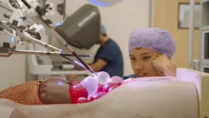 Een foto van een persoon die zit te kijken naar een robot die een plastic pop opereert. De robot bestaat uit 3 lange metalen stangen met klemmen en lampjes. De stangen grijpen kleine ballonnen vast die in de open buik van de pop liggen.