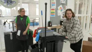 Een foto van presentator Pascal die een schoonmaakwagen duwt. Er staat een schoonmaker naast hem die naar hem kijkt.