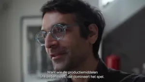 Een foto van een man die voor de camera staat te praten. Er is een ondertitel in beeld met de tekst 'want wie de productiemiddelen in handen heeft, die domineert het spel'.