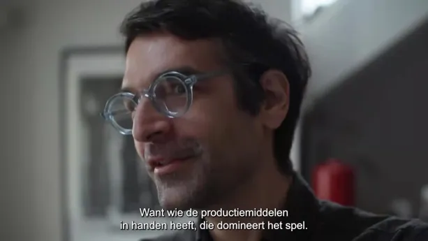Een foto van een man die voor de camera staat te praten. Er is een ondertitel in beeld met de tekst 'want wie de productiemiddelen in handen heeft, die domineert het spel'.
