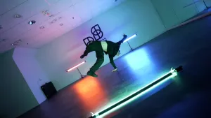 Een schuine foto van een breakdancer die een draai maakt op een hand. De dansruimte wordt verlicht met een paar felle lichtbalken in verschillende kleuren. Achter op een muur staat een logo.