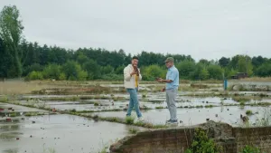 Een foto van twee mensen die buiten staan te praten. Ze staan op betonnen platen met onkruid ertussen. Ze hebben stenen in hun handen.