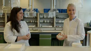 Een foto van presentator Janouk en een andere persoon, die achter een tafel staan. Ze hebben witte werkkleding aan en staan in een ruimte met heel veel potjes en doosjes. Janouk heeft een kommetje vast.