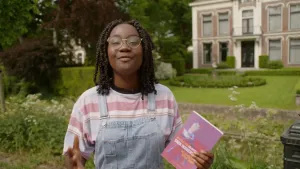 Een foto van presentator Tirsa met een boek in haar hand. Het boek heet "Historie van mejuffrouw Sara Burgerhart". Ze staat voor een deftig huis met een tuin.