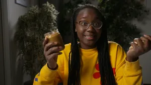 Een foto van presentator Tirsa met een open pot pindakaas in haar rechterhand en een aantal pinda's in haar linkerhand.