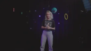 Een foto van presentator Janouk die in het donker staat voor een gordijn waar sterren, ringen en een planeet op geschenen wordt.