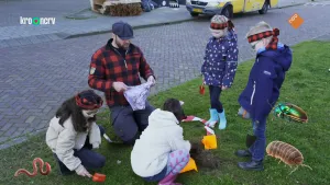 Een foto van boswachter Tim en vier kinderen op een grasveldje bij een straat. Twee kinderen staan te kijken. Tim en de twee anderen zitten op de grond met een zakje en schepjes. Op de foto zijn foto's geplakt van een regenworm, een pissebed en een kever.