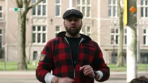 Een foto van boswachter Tim in een straat. Achter hem is een straat te zien met bomen ernaast. Daarachter is een groot gebouw. Tim kijkt en praat naar de camera.