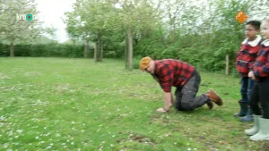 Een foto van boswachter Tim en twee kinderen op een grasveld met bomen en struiken eromheen. De twee kinderen staan helemaal rechts op de foto. Iets meer naar het midden zit Tim op handen en knieën op het gras.